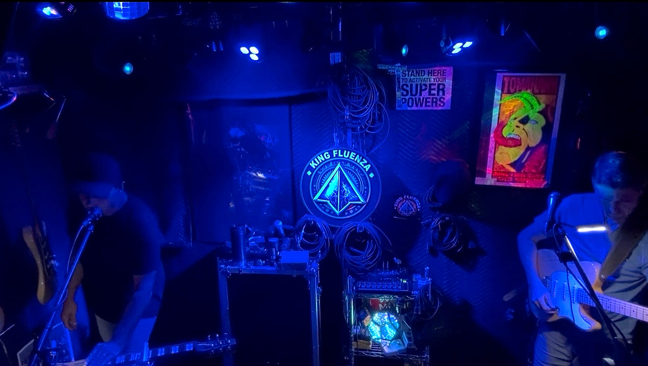 Band running a song under blue wash lighting beside the King Fluenza banner